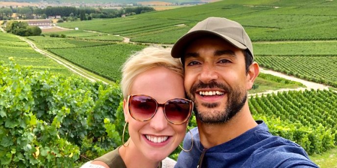 A smiling couple taking a selfie while standing at the edge or rolling vineyards in the Champagne region