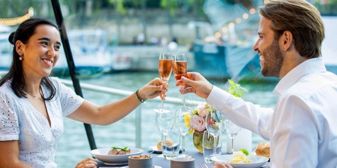 A couple toasting with wine glasses during a romantic paris dinner cruise on the Seine River, with scenic views of historic Parisian buildings and trees under a blue sky.