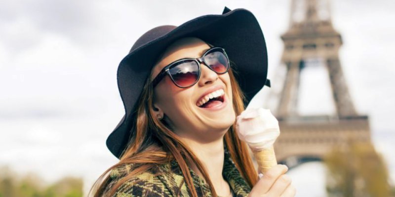 At the Eiffel Tower a young woman with a hat and sunglasses enjoys an ice cream cone